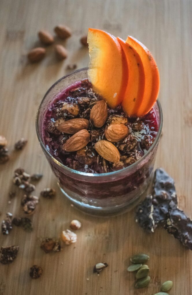 A layered chia pudding served in a glass with chocolate chia base, raspberry layer, topped with peach slices, almonds, granola, and seeds on a wooden surface.