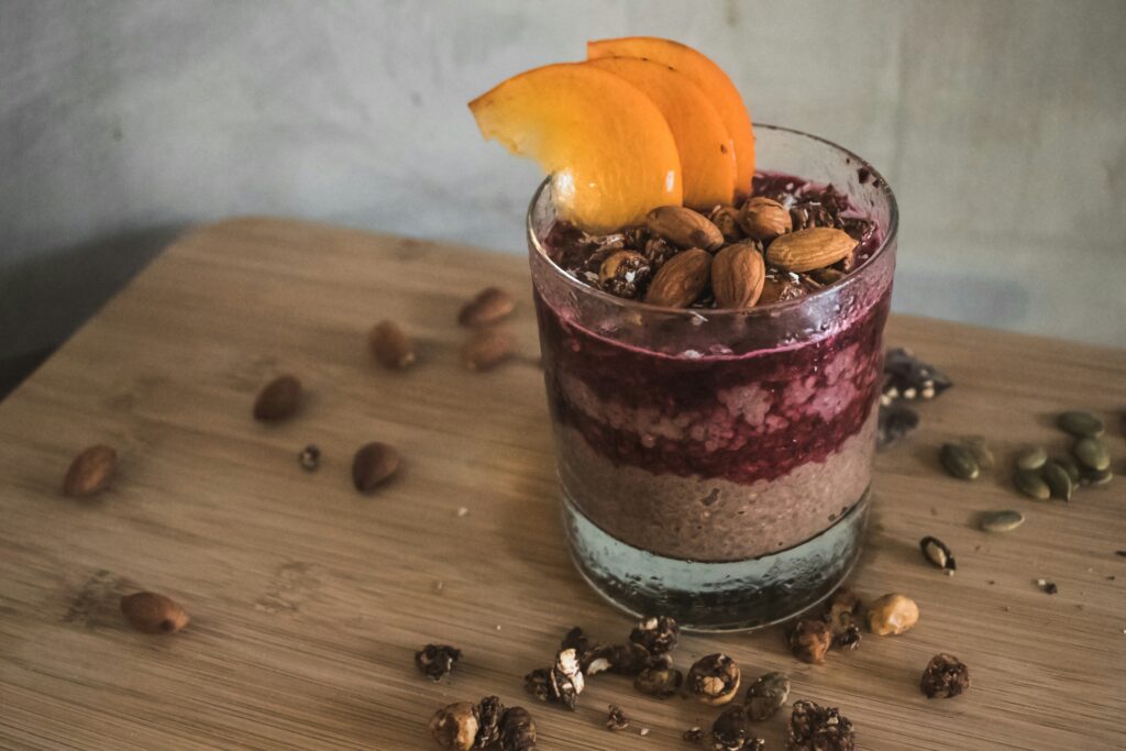 A layered chia pudding served in a glass with chocolate chia base, raspberry layer, topped with peach slices, almonds, granola, and seeds on a wooden surface.