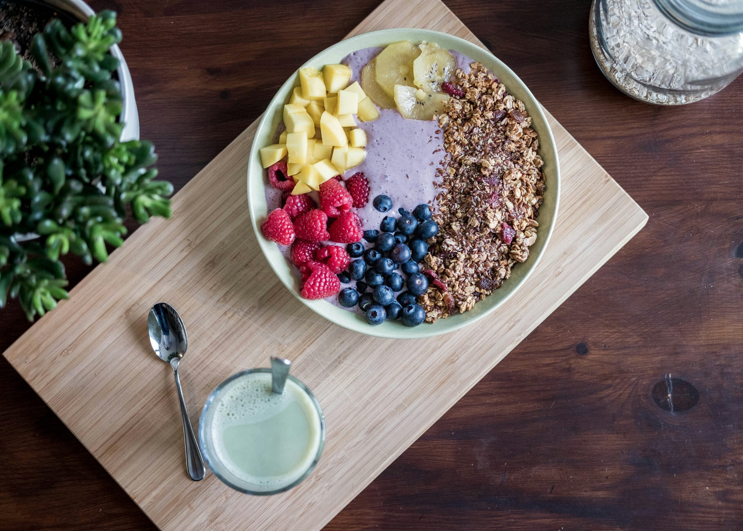 A colorful berry smoothie bowl topped with fresh raspberries, blueberries, diced mango, kiwi slices, and crunchy granola on a wooden board with a glass of green matcha latte on the side