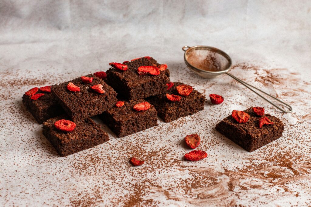 Healthy chocolate strawberry brownies topped with dried strawberry slices, placed on a white surface dusted with cocoa powder alongside a metal sieve.