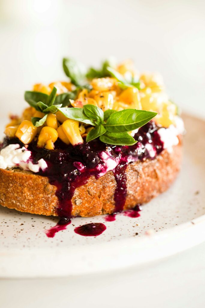 A plate of healthy corn and berry toast featuring toasted bread topped with creamy ricotta, vibrant blueberries, roasted corn kernels, fresh basil leaves, and a drizzle of berry compote, served on a white speckled plate.