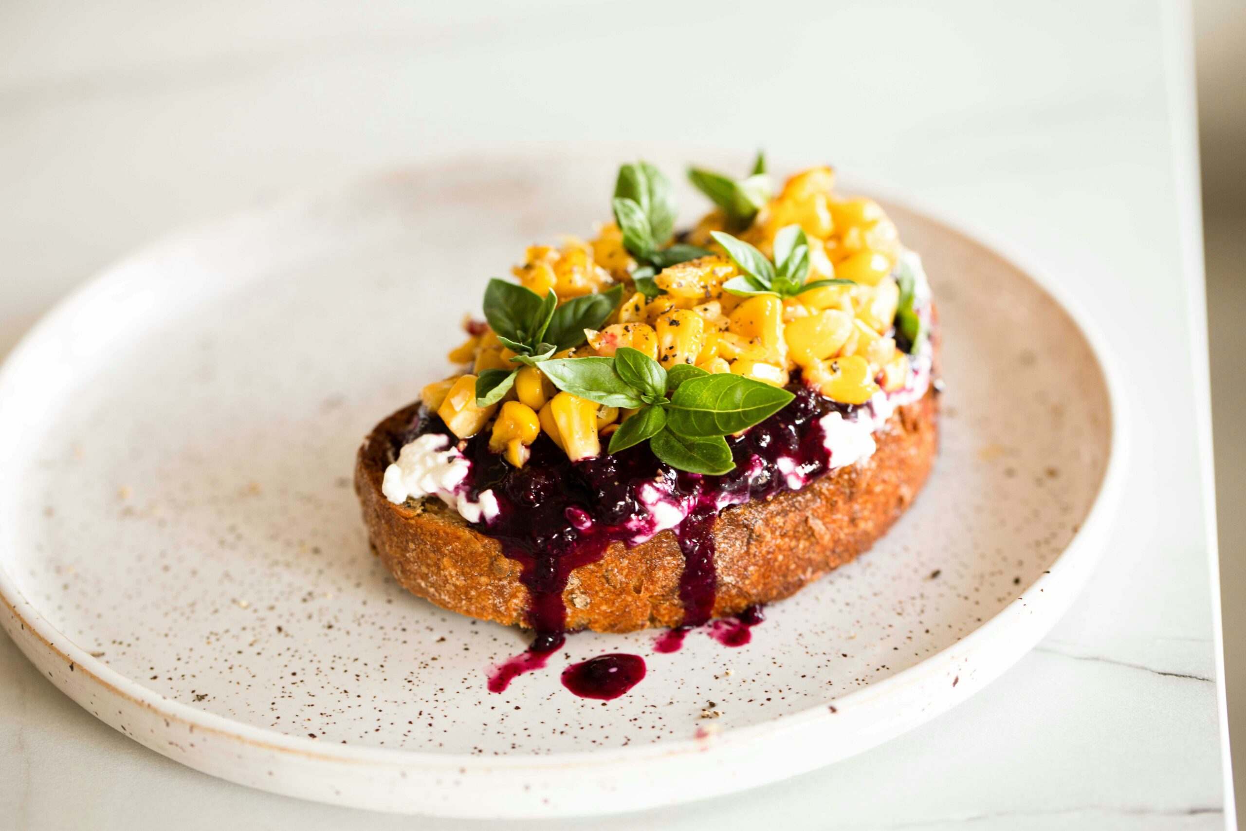 A plate of healthy corn and berry toast featuring toasted bread topped with creamy ricotta, vibrant blueberries, roasted corn kernels, fresh basil leaves, and a drizzle of berry compote, served on a white speckled plate.