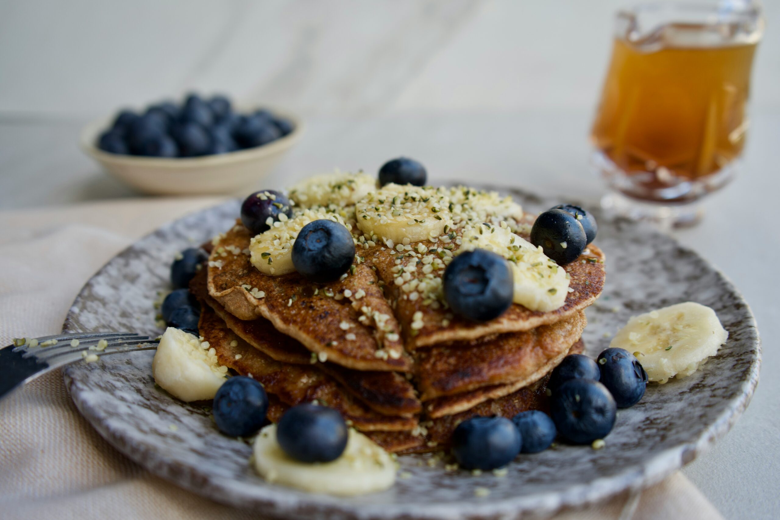 Stack of healthy pancakes topped with fresh blueberries, banana slices, and hemp seeds, served on a rustic plate with maple syrup in the background.