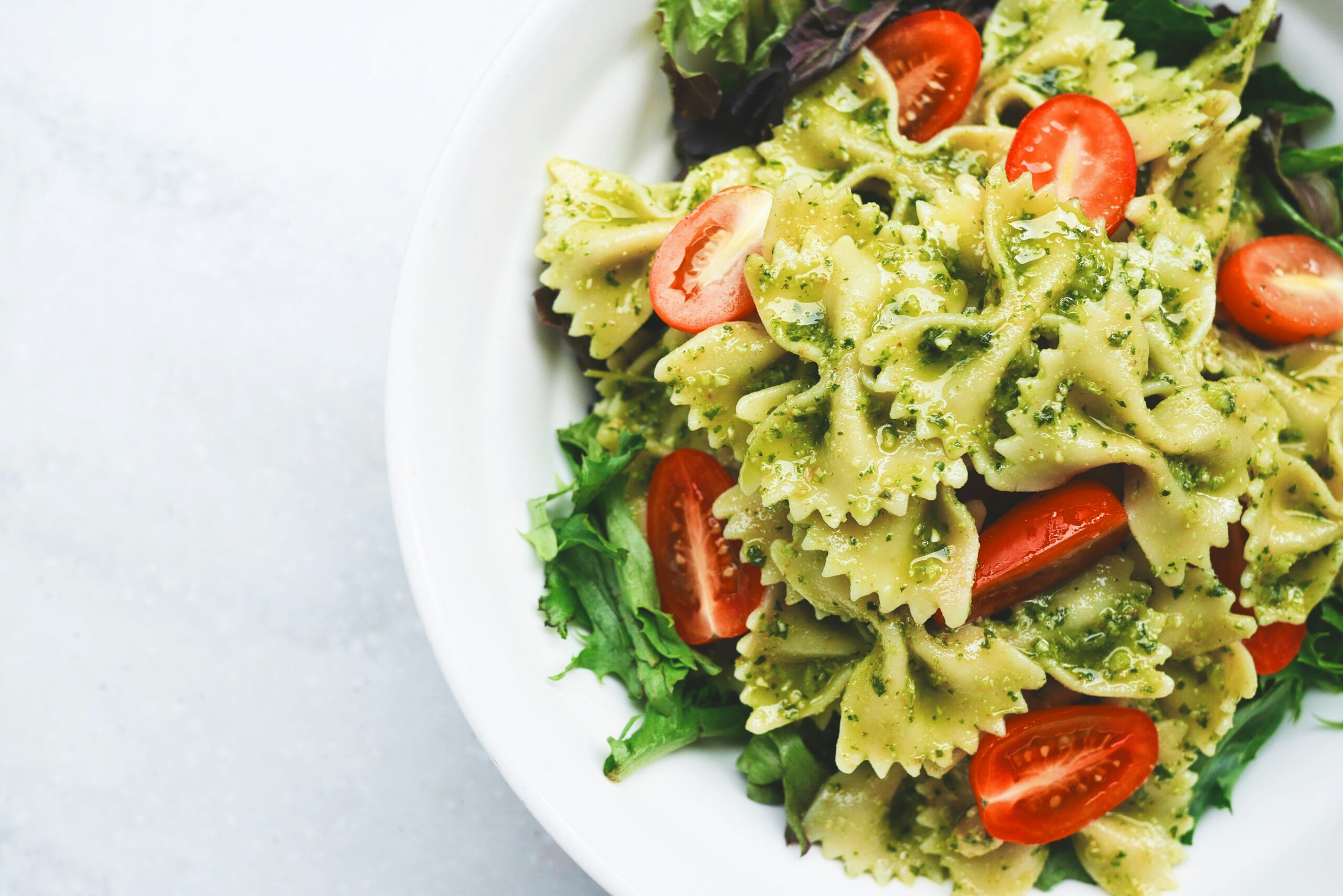 A close-up of a vibrant pesto pasta salad made with bowtie pasta, cherry tomatoes, and fresh mixed greens, served in a white bowl on a light background.