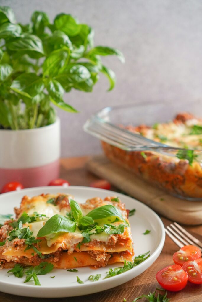 A plate of healthy lasagna topped with fresh basil leaves, served on a wooden table with cherry tomatoes and a glass baking dish of lasagna in the background.