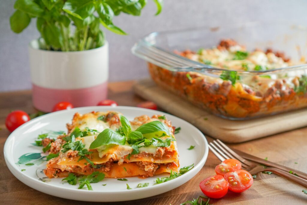 A plate of healthy lasagna topped with fresh basil leaves, served on a wooden table with cherry tomatoes and a glass baking dish of lasagna in the background.