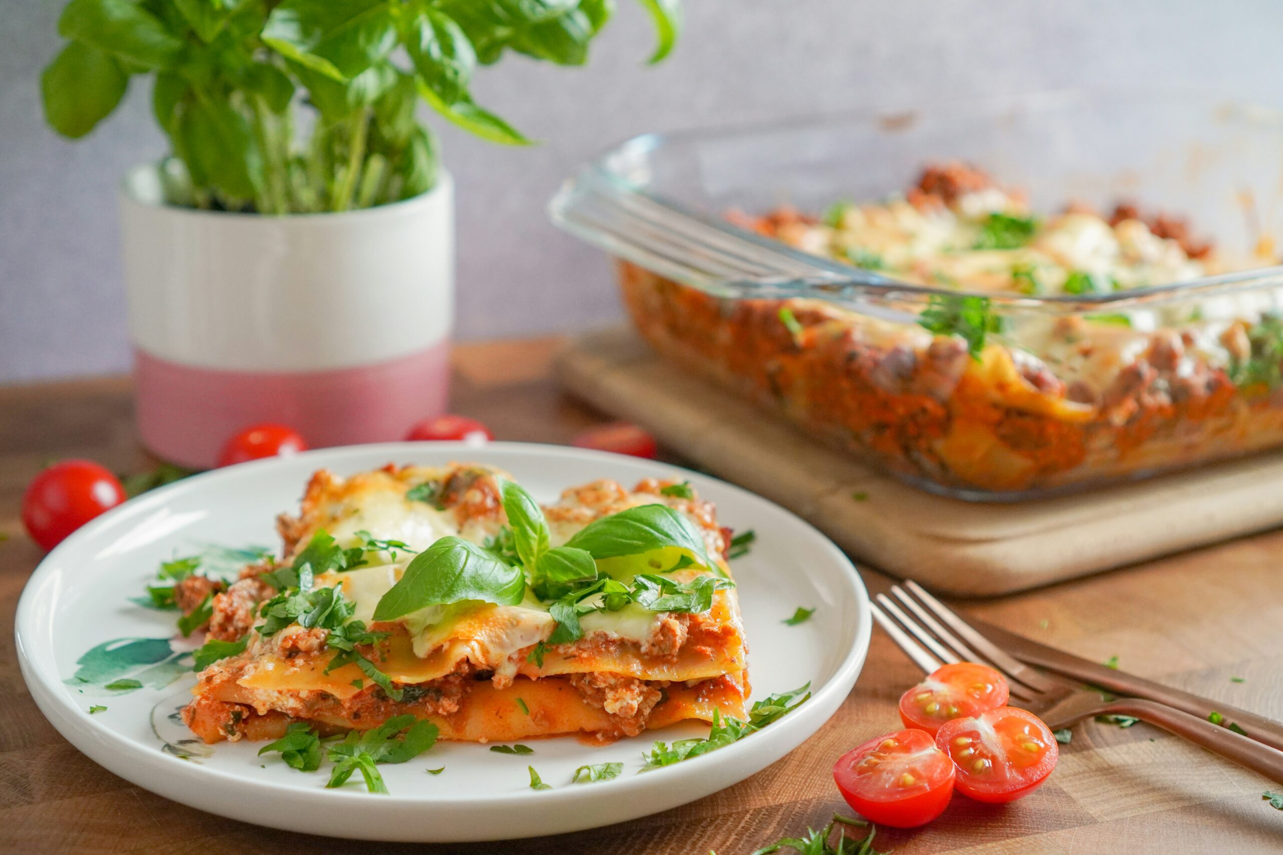 A plate of healthy lasagna topped with fresh basil leaves, served on a wooden table with cherry tomatoes and a glass baking dish of lasagna in the background.