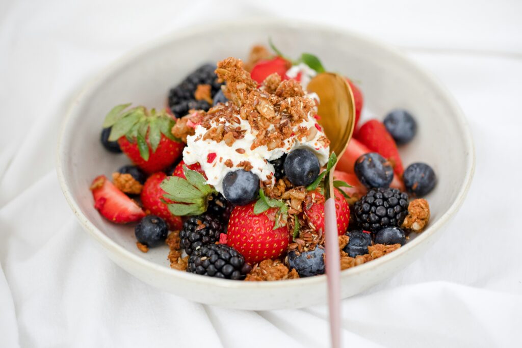 A bowl of fresh mixed berries, including strawberries, blueberries, and blackberries, topped with whipped cream, crunchy granola, and a golden spoon, served on a white background.