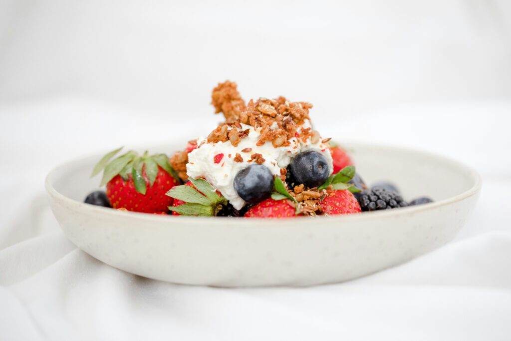 A bowl of fresh mixed berries, including strawberries, blueberries, and blackberries, topped with whipped cream, crunchy granola, and a golden spoon, served on a white background.